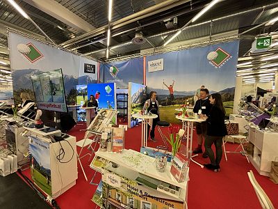 Exhibition booth with golf theme, featuring displays, banners, and visitors interacting on a red-carpeted floor under bright overhead lights.