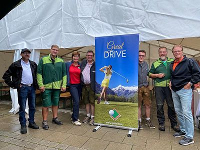 Seven people stand in front of a tent with a golf-themed banner, smiling and posing for a group photo at an outdoor event.