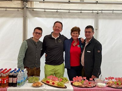 Vier Personen stehen in einem Festzelt hinter einem Tisch mit Getränken und Snacks. Sie lächeln freundlich in die Kamera. Der Tisch ist mit kariertem Tuch bedeckt.