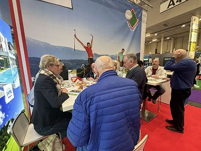 People seated at a table having a discussion at a trade show booth, with a scenic backdrop and two standing figures in business attire.