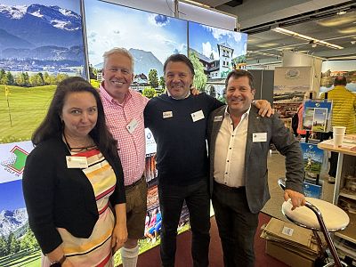 Vier Personen stehen lächelnd vor einem Stand auf einer Messe, umgeben von Landschaftsbildern. Drei Männer und eine Frau tragen Namensschilder.