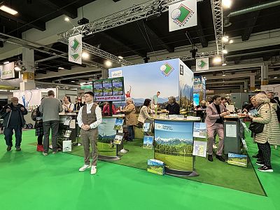 People gather at a colorful trade fair booth with scenic displays. Representatives provide information and brochures. The space has a vibrant and inviting setup.