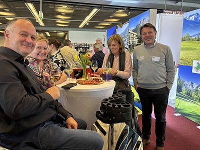 People at a networking event enjoying drinks and snacks around a high table, with exhibition stands and a golf bag in the setting, creating a casual vibe.