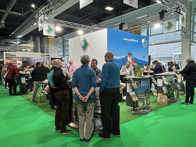 People gather around a trade fair booth with brochures and travel displays. The green floor and banners suggest a tourism or travel industry event.