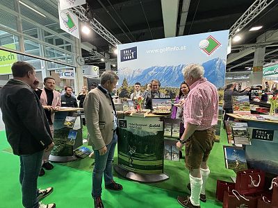 People interact at a trade fair booth displaying scenic landscapes. The setting is busy with attendees in casual attire, discussing and exploring the exhibits.