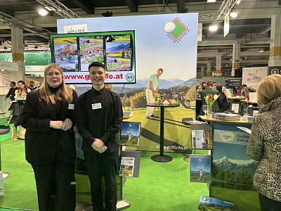 Two people at a brightly lit booth at a tourism fair with scenic posters and interactive screens displaying outdoor destinations.