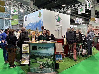 People gather at a trade show booth with mountain images, exploring brochures and products. The setting is vibrant with social interactions and green flooring.
