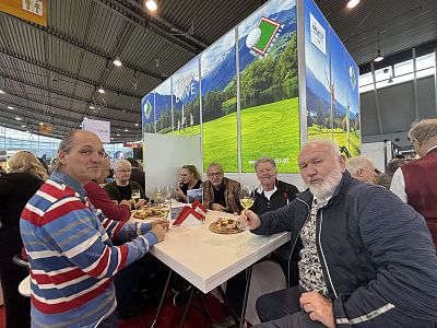 People sitting around a table eating at a trade show, with a backdrop displaying scenic landscapes. The scene is lively and social.