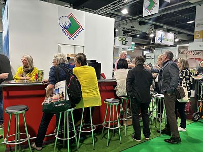 People gathered at a bright exhibition booth with golf-themed decorations, some standing and others sitting on stools, engaging in lively discussions.