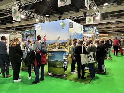 People gather at a vibrant travel fair booth with mountain images and display materials, engaging with representatives. Bright green flooring adds to the lively atmosphere.