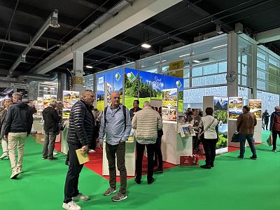 People engage with exhibitors at a travel expo, featuring colorful displays of tourist destinations. The venue is lively with visitors exploring various booths.