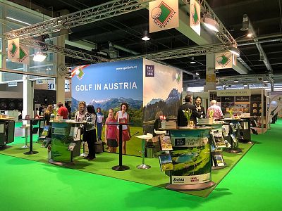 A trade show booth displaying 'Golf in Austria,' with posters of mountains and people interacting. A green carpet complements the natural theme.