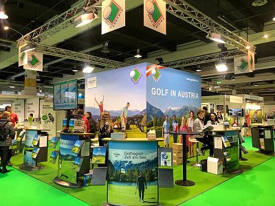 A vibrant exhibition booth at a travel expo promotes Austrian tourism, featuring banners with mountain landscapes and golf. Visitors engage with the displays.