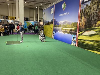 Indoor golf demo with audience, featuring a presenter and a backdrop of golf course images.
