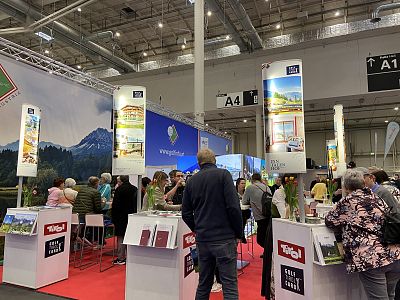 People at a tourism trade fair with brochures and posters, surrounded by display stands and a large backdrop of mountains in an exhibition hall.
