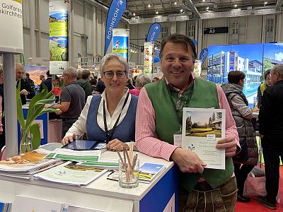 Zwei Personen in traditioneller Kleidung stehen lächelnd an einem Infostand auf einer Messe, halten Flyer in den Händen. Im Hintergrund sieht man Displays.