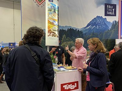 Several people at an exhibition booth enjoy conversation and snacks, with a mountain backdrop, suggesting a tourism or travel theme.