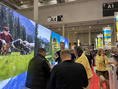 Attendees gather at a convention booth displaying scenic mountain images, with people in conversation. Large scenic panels and a red carpet are visible.