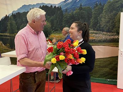 A man and a woman smiling at each other while holding a bouquet of red and yellow tulips. The background shows a scenic view of trees and mountains.