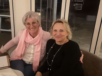 Zwei lächelnde Frauen sitzen nah beieinander in einem Innenraum, eine trägt eine rosa Strickjacke und Schal, die andere einen schwarzen Pullover.
