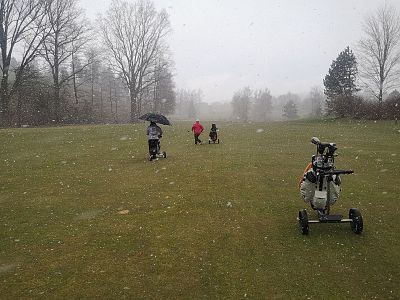 Golfers on a snowy course, trees without leaves. One person with an umbrella. Golf bags on carts, overcast sky suggests a cold, wintry afternoon.