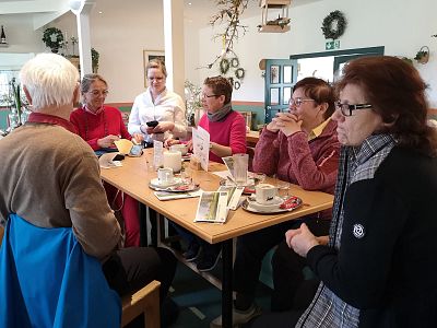 Eine Gruppe von sechs älteren Menschen sitzt an einem Tisch in einem gemütlichen Café. Sie unterhalten sich bei Kaffee und Kuchen in entspannter Atmosphäre.