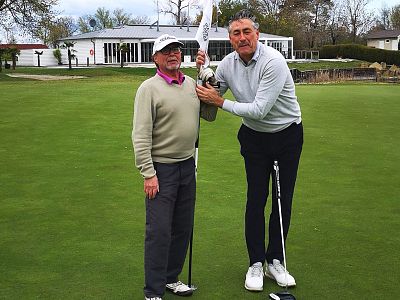 Zwei ältere Männer stehen auf einem Golfplatz. Einer hält einen Golfschläger, der andere eine Flagge. Im Hintergrund ist ein Clubhaus zu sehen.