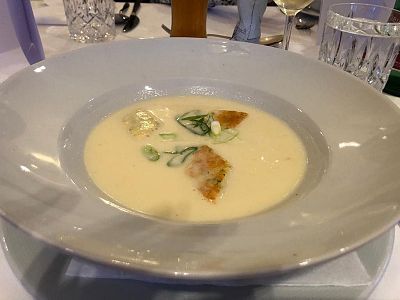 A bowl of creamy soup with garnish sits on a dining table, surrounded by glasses and tableware.