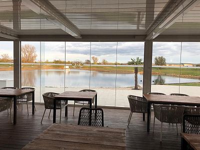 Indoor seating area with wooden tables and chairs, large windows showing a tranquil lake and trees, creating a peaceful atmosphere.