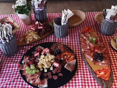A table with red and white checkered cloth displaying plates of assorted cheeses, cured meats, tomatoes, grapes, and decorative items.