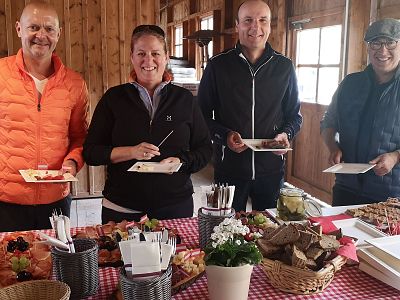 Four people stand smiling with plates of food in a rustic setting. The table is covered with a red checkered cloth and various dishes, including bread and salad.