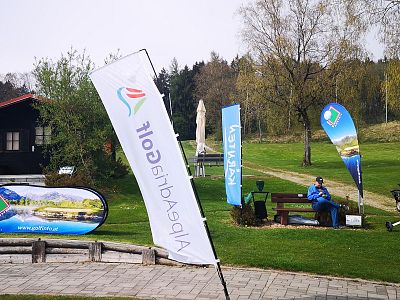 Grass area with several promotional banners and a person sitting on a bench surrounded by trees and a path.