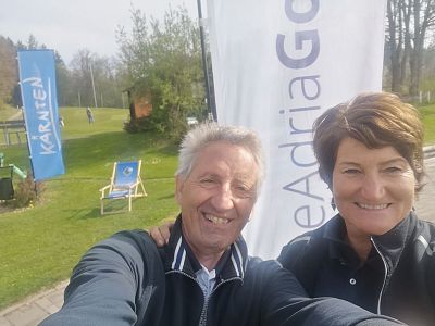 A couple takes a selfie on a golf course with flags in the background. It's a sunny day, and the area is surrounded by green grass and trees.