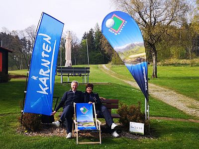 Two people sit on a bench between Carinthia banners in a green park area, enjoying a sunny day with promotional flags around them, suggesting a leisure event.