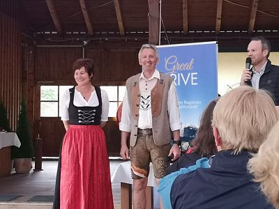 Three people at an indoor event. Two are wearing traditional German attire, while one is speaking into a microphone. The setting seems festive.
