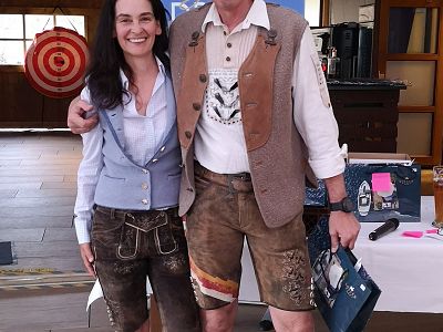 A smiling man and woman in Bavarian attire stand indoors, wearing lederhosen and dirndl, symbolizing a cultural or festive occasion.