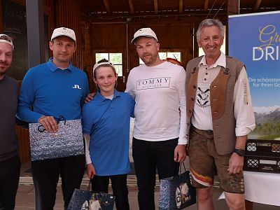 Gruppe von fünf Personen in Freizeitkleidung, einige mit Baseballkappen, posiert für ein Foto. Dahinter ein Holzgebäude und ein Banner mit der Aufschrift "Great Drives".