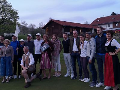 A group of people pose for a photo on a golf course. Some are in traditional Bavarian attire, while others wear casual or athletic clothing.