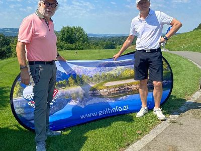 Two men stand on a golf course holding a colorful banner, with a scenic landscape in the background.
