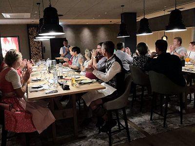 A group of people in traditional outfits seated at a long dinner table in a warmly lit room, creating a festive and cozy atmosphere.