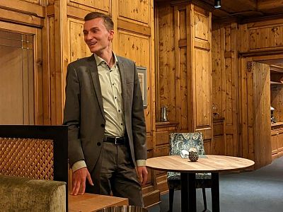 A man in a gray suit stands in a warmly decorated room with wooden walls and round tables, smiling and looking to the side.