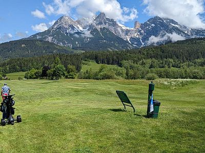 Lush green golf course with a golf bag on the grass, surrounded by tall pine trees and majestic mountains in the background under a partly cloudy blue sky.
