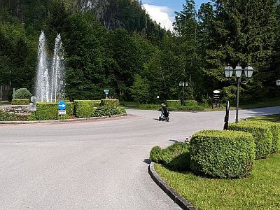 A park with a large fountain surrounded by trees and bushes. A person on a bicycle rides on a path in the foreground on a sunny day.
