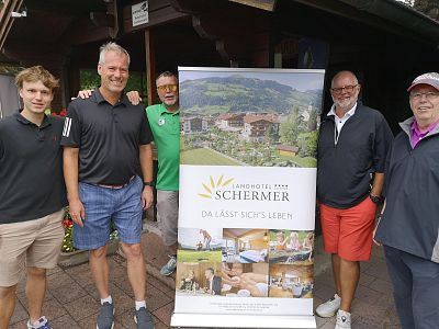 Fünf Männer stehen lächelnd im Freien neben einem Werbebanner des Landhotels Schermer, das eine idyllische Landschaft und Hotelbilder zeigt.