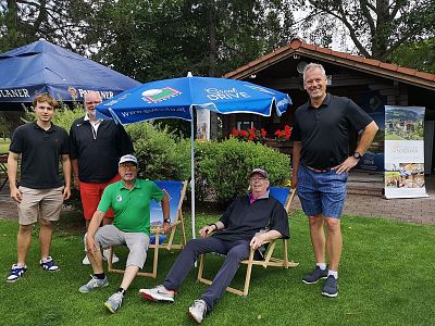 Five adults at a golf course, with two sitting under an umbrella and three standing, all smiling. A clubhouse and trees are visible in the background.