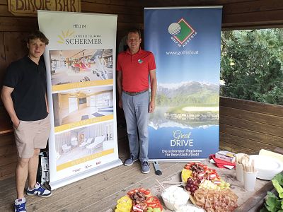 Zwei Männer stehen in einem Holzpavillon neben Werbebannern. Ein Tisch mit einer Auswahl an Lebensmitteln steht im Vordergrund. Gartenumgebung im Hintergrund.