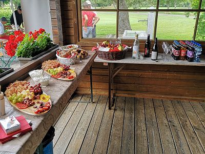 A buffet with fruits and cheeses on wooden tables, decorated with red flowers, set on a wooden terrace overlooking a garden.