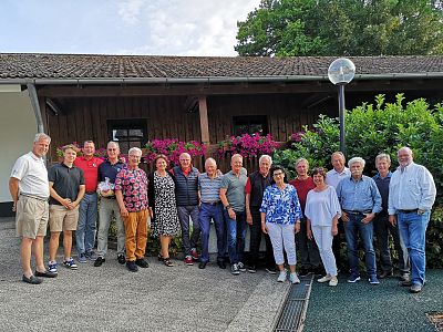 A diverse group of men and women stand together for a photo in front of a wooden building with pink flowers. The scene is bright, suggesting a daytime gathering.