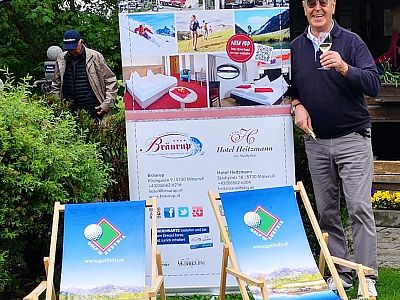 A man stands next to two branded chairs and a hotel advertisement banner outdoors, holding a drink, in a garden setting with another person in the background.