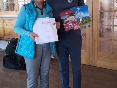 Zwei Personen stehen nebeneinander in einem Raum mit Holzwänden. Eine Person hält ein Blatt Papier und die andere hält zwei bunte Broschüren. Beide lächeln.